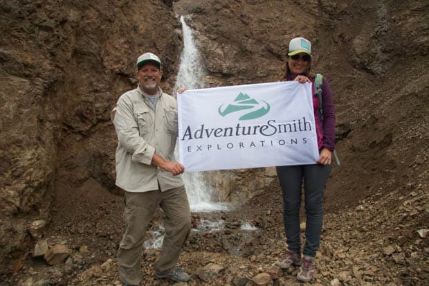 A man and a woman holding a banner in front of a waterfall in Alaska. 