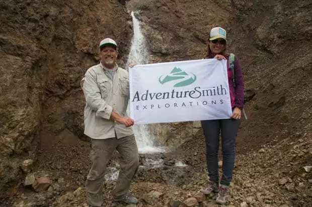 A man and a woman holding a banner in front of a waterfall in Alaska. 