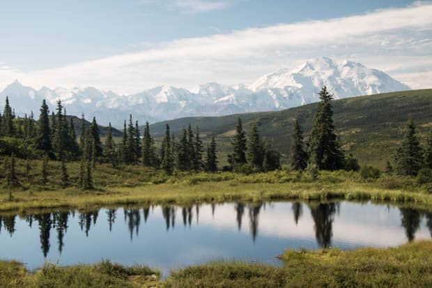 A pool of water with trees and snow covered mountain tops in the distance. 