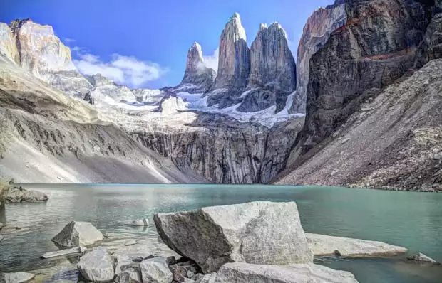 Landscape of a glaciated teal blue lake with sheer rock mountains and granite towers with snow on Patagonia land tour Towers Base hike.