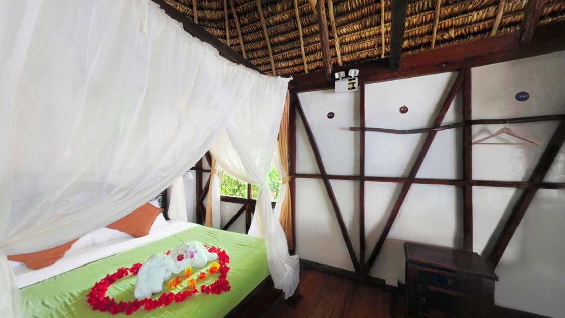 Large bed with green bedspread, white sheets & white canopy looks up at a thatched roof.