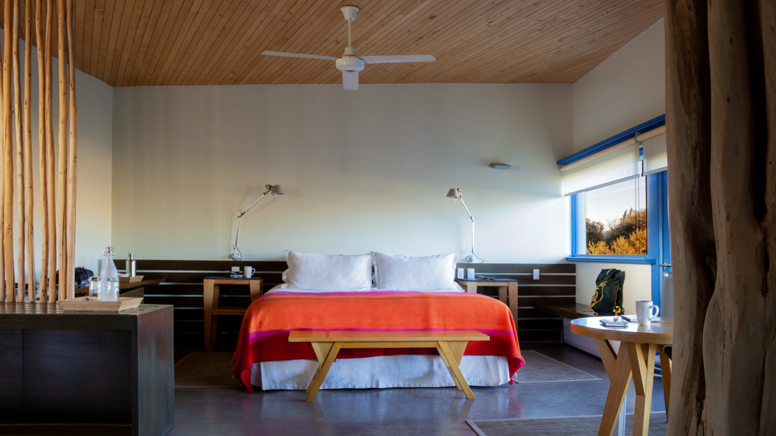 King bed with white linens & orange blanket, ceiling fan, a small table & large view windows in Catur Suite at Explora Atacama.