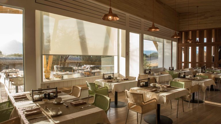 Indoor dining room at Explora Atacama with large view windows, wicker chairs, 2-top tables set for dinner & earth-tone decor.