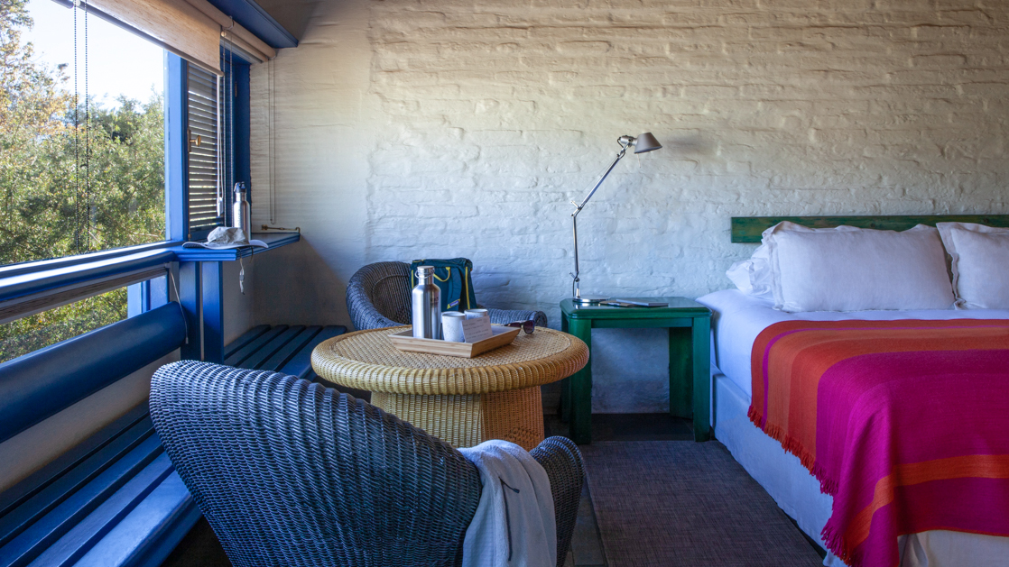 Double bed, green beside table, small wicker table & 2 chairs by view window in Yali Room at Explora Atacama Lodge in Chile.