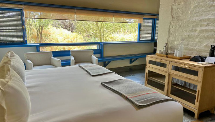 Tulur Room at Explora Hotel Atacama with queen bed in white linens, small wood table, large window, white walls & blue accents.