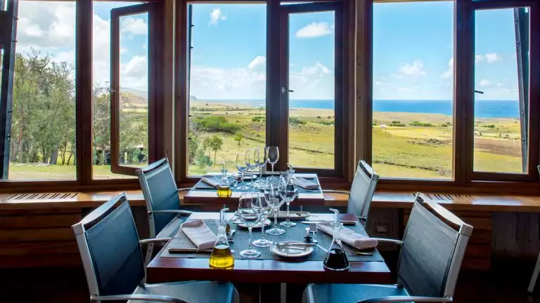 Two 2-top tables with modern chairs are set for a fancy dinner by view windows looking out onto rolling grassy hills & ocean.