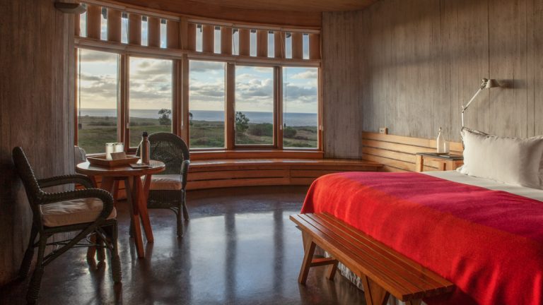 Large double bed with red blanket plus small wicker chairs & table in wood & stone room with large view windows overlooking sea.