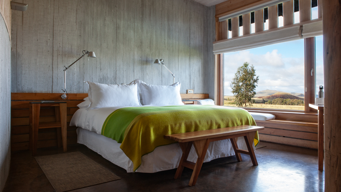 Double bed with white sheets & green blanket in stone & wood room with view window at Explora Rapa Nui.