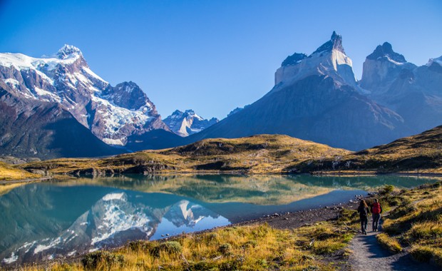 2 Patagonia travelers walking on a trail next to a lake reflecting the snow capped mountains in Torres del Paine National Park.