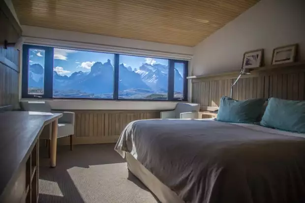 Explora Lodge view of a room with a bed, chairs, desk and a window with a panoramic view of mountains in Torres del Paine National Park land tour.