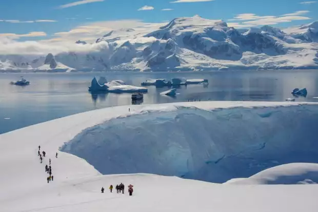 People snowshoeing uphill on Antarctica with their small cruise ship in the background. 