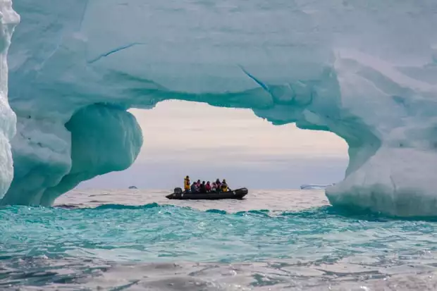 Guest from a small ship on a skiff under the arch of an iceberg.