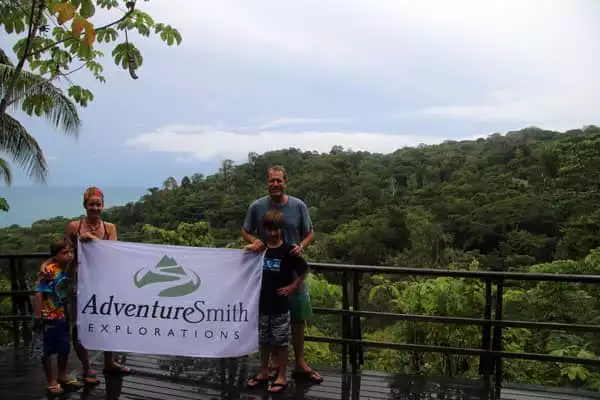 Family of travelers in Costa Rica holding a Adventuresmith Explorations flag on a deck in the rainforest with the ocean in the background.