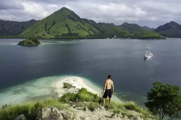 A man in boardshorts and no shirt stands on a hilltop above a small sandy beach peninsula with no one on it and a small ship and volcanoes in the water below