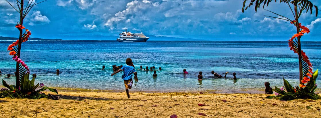 Local children playing on the beach and in the ocean with a small cruise ship in the background. 