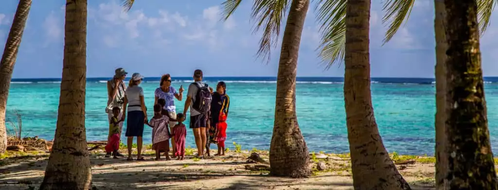 Travelers stand hand in hand with children villagers between five large oceanfront palm trees