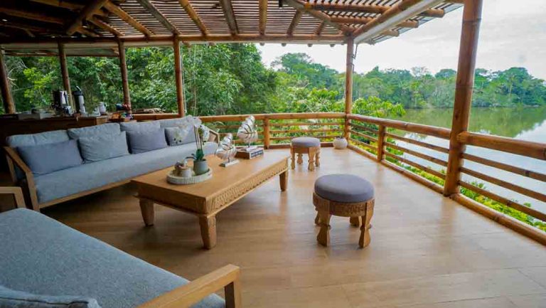 Open-air lounge at La Selva Ecuador with thatch roof, wood floor & coffee table & padded blue couches beside a lake.