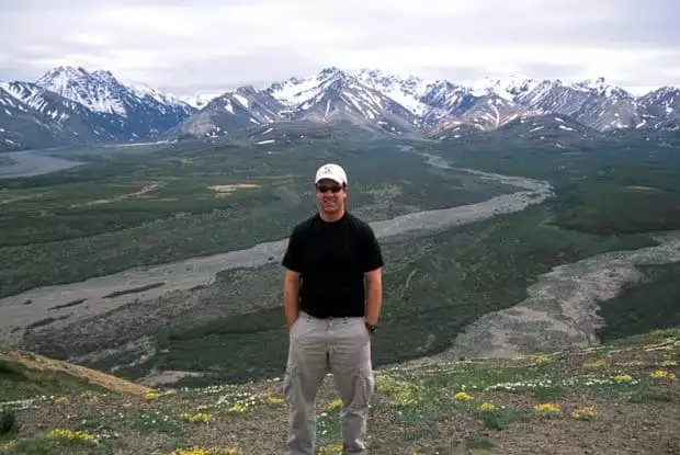 Alaskan traveler posing on top of a mountainside with wildflowers, wild muddy rivers and snow capped mountains.