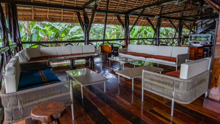 Open-air lounge at Napo Wildlife Center with dark wood floors, thatched roof & padded white couches with colorful woven blankets.