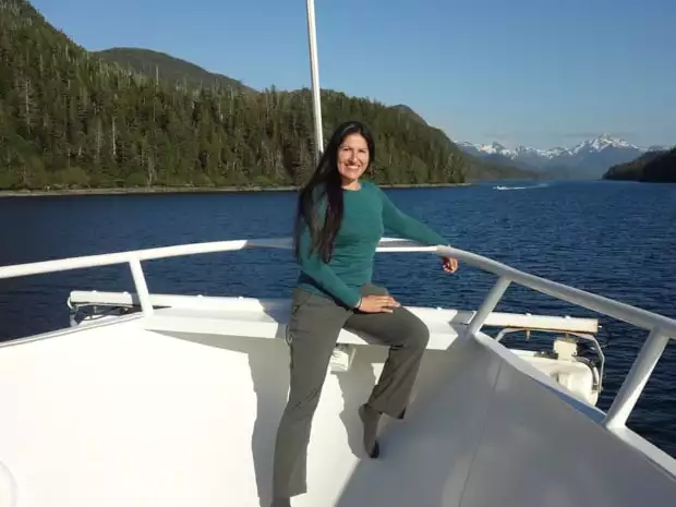 Alaskan traveler posing on the deck of a small ship cruise with snowcapped mountains and forest in the background.