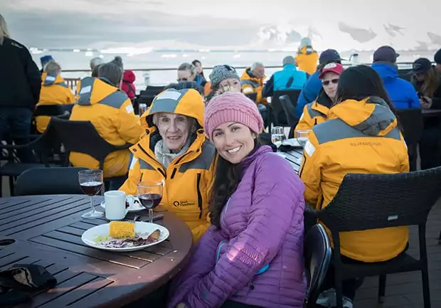 Guests aboard small ship in Antarctica enjoy a bbq on the deck of the ship and drinking wine with icebergs in the background. 