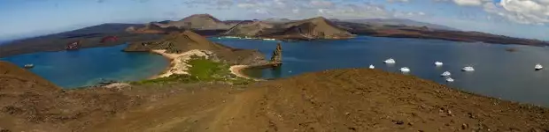 Panoramic view of Bartolome Island with small boats and yachts anchored, rock formations, a hourglass sandy shoreline.