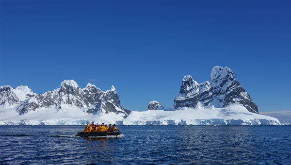 Travelers in a skiff on the ocean pass in front of white-capped Antarctica mountain