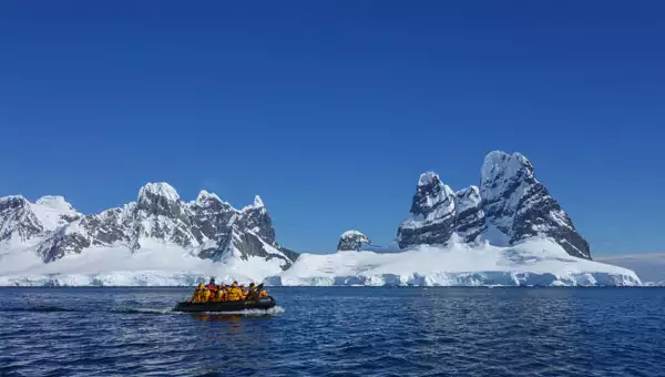 Travelers in a skiff on the ocean pass in front of white-capped Antarctica mountain