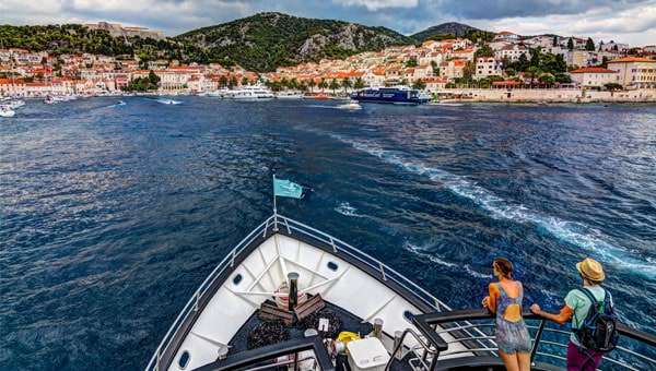Guests aboard a small Mediterranean yacht approach a hilly shoreline with terra-cotta-roofed buildings.