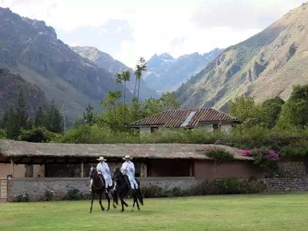 Hotel in a valley floor with large mountain ranges on either side and 2 cowboys riding horses on a lawn.