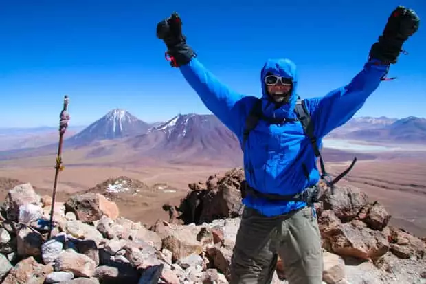 Author on the top of a mountain with arms in the air in celebration of summitting with mountains and plains in the background.