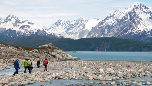 Alaska travelers hike on a rocky shoreline with ocean and snowy mountains