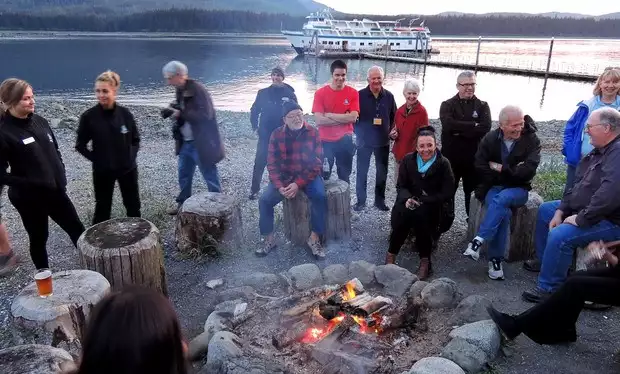 Guests at Orca Point Lodge in Alaska sitting around a campfire on the beach with their small ship in the background. 