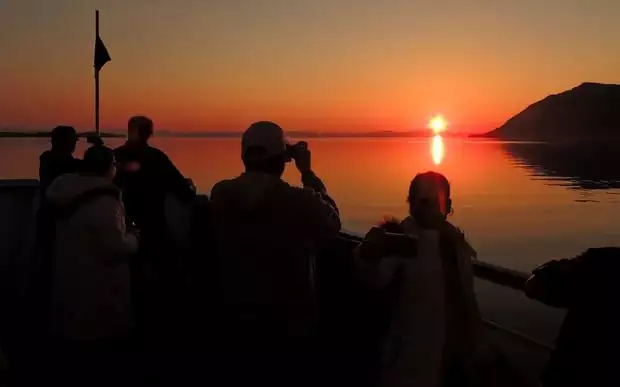 Guests on the bow of their small ship cruise in Alaska taking a picture of the orange sunset. 