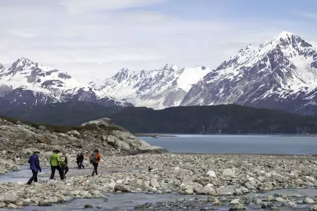 Group of Alaskan travelers hiking on a rocky shoreline with snowy mountains and small ship cruise in the background.