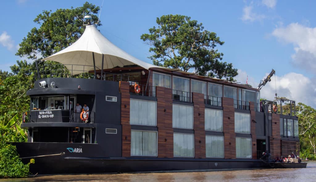 The outside of the Aria luxury riverboat while sailing through the Amazon. Big windows on every floor, sundeck with large sun unbrella and a skiff full of people.