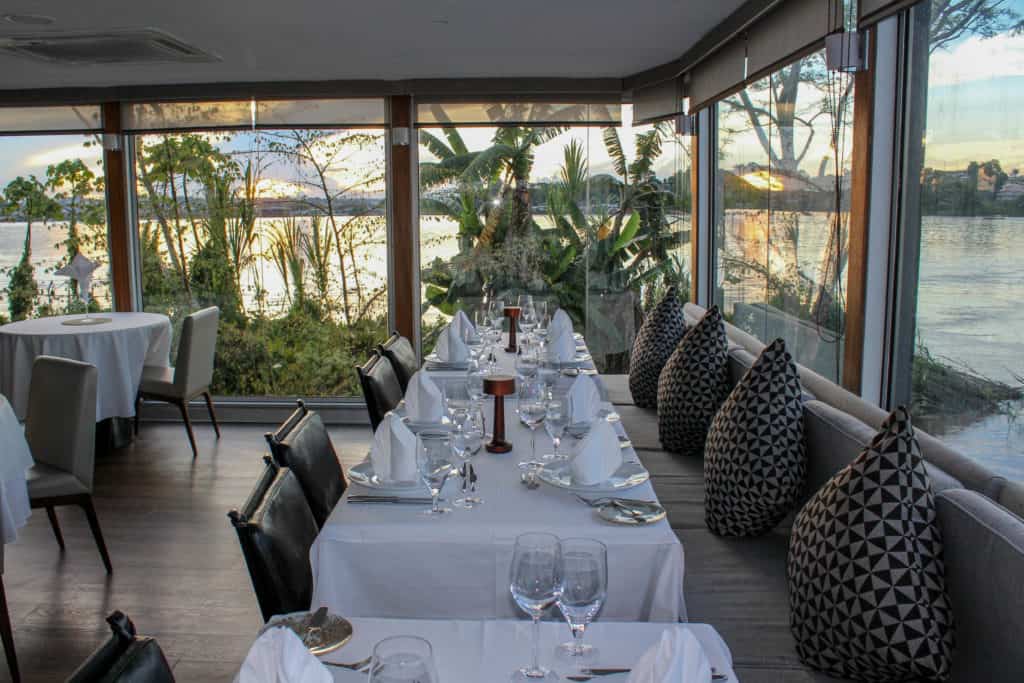 Dining area aboard the Aria with white table clothes, large windows and the sunsetting out outside.