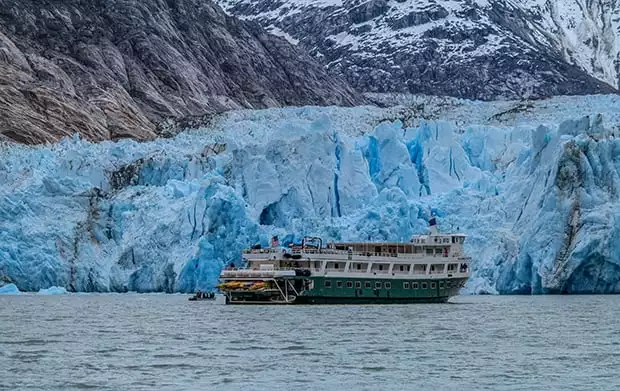 The Wilderness Discoverer small Alaska cruise ship with a green hull sits close to a big blue glacier.
