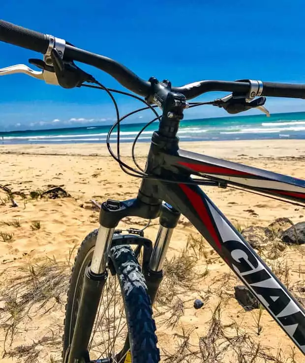 A mountain bike on a sandy beach with waves in the ocean on a sunny day.