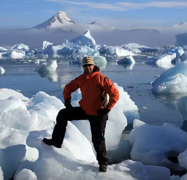 Guest aboard a small ship cruise in Antarctica posing with foot on top of an iceberg near shore with many icebergs in the background. 