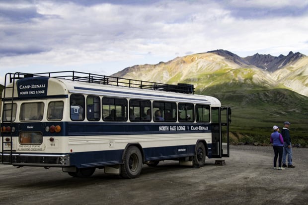 Two travelers visiting Denali National Park stand beside their Camp Denali private tour bus with mountains behind them.