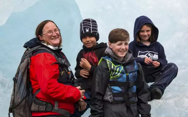 A woman guide in Alaska with a group of smiling kids sitting on an iceberg.