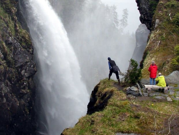 Three people standing near a cliff edge across from a raging waterfall in Alaska. 