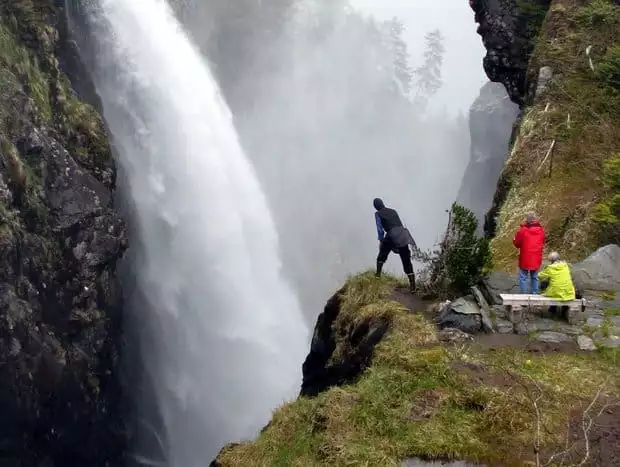 Three people standing near a cliff edge across from a raging waterfall in Alaska. 