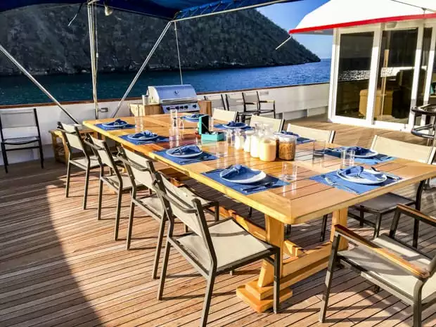 A guest table set for dining on the deck of the small ship cruise Passion on a sunny day while anchored off the coastline in the Galapagos.