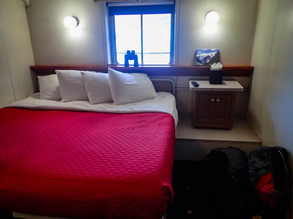 inside a cabin aboard Wilderness Adventurer, a red comforter wraps the bed with white pillows, on the wall is a sliding glass window with binoculars on the window sill. 