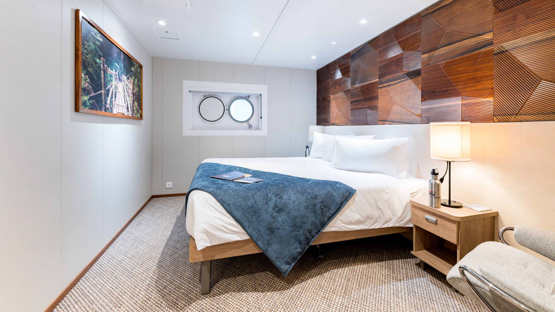 Coral Deck Stateroom with junior king bed, single porthole, framed photograph on wall, wooden nightstand with lamp & metal water bottle, and side chair aboard Coral Geographer.