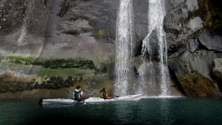 Two kayakers paddling past twin waterfalls flowing down high rock walls in Alaska.