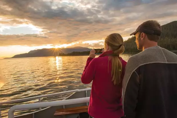 On a San Juan Islands cruise two guests stand onboard with a camera facing the sun setting over an island in the distance.
