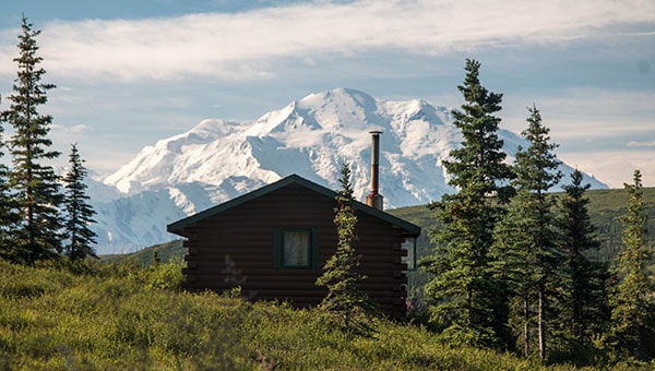 A small Denali National Park log cabin with the snowy Denali peak behind it